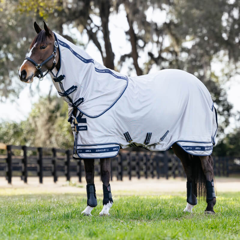 A horse wears the LeMieux Arika Armour-Tek Fly Rug Grey, standing on grass with trees behind.