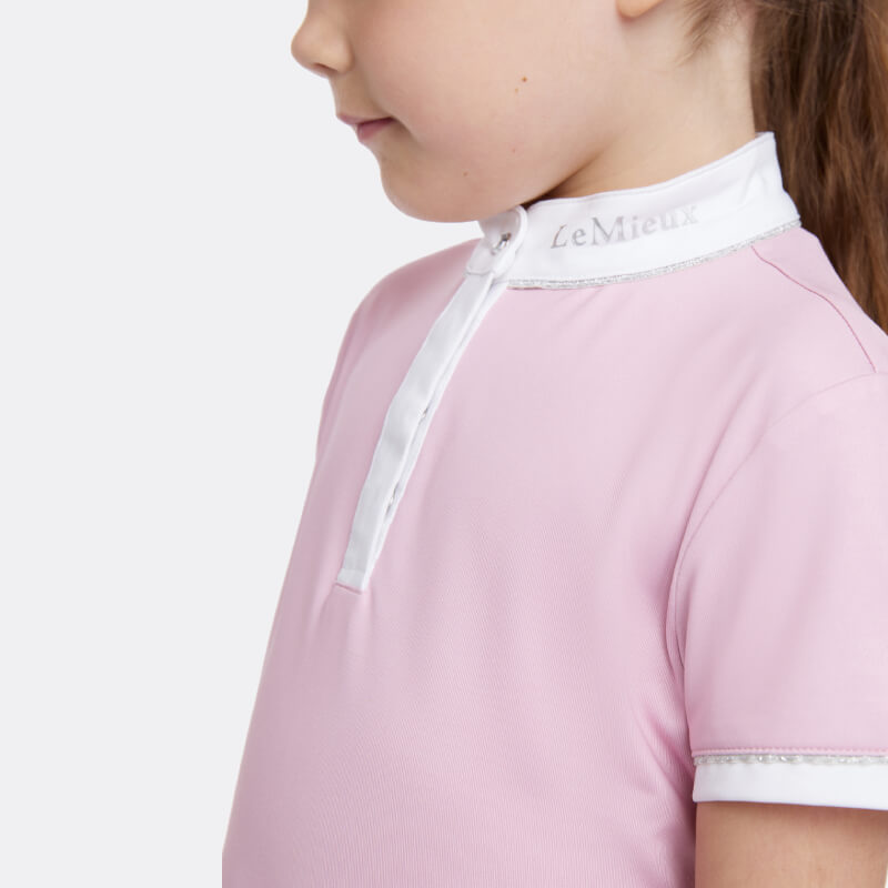 Smiling girl in a LeMieux Mini Bella Show Shirt Blossom and beige pants stands against white background.