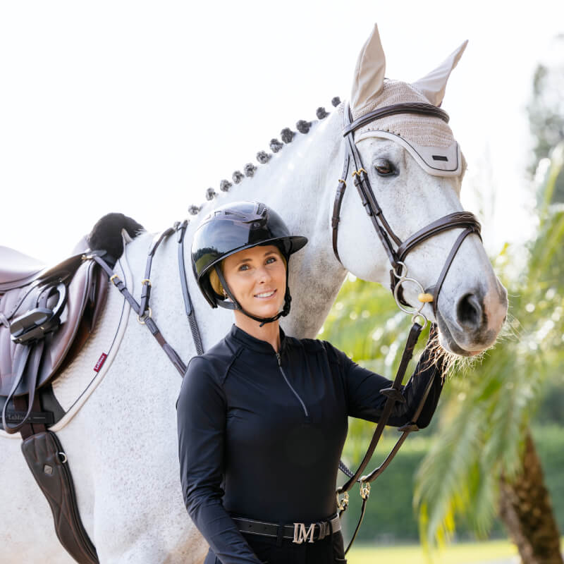 Smiling equestrian woman in helmet stands beside a white horse in LeMieux Essence Acoustic Fly Hood.