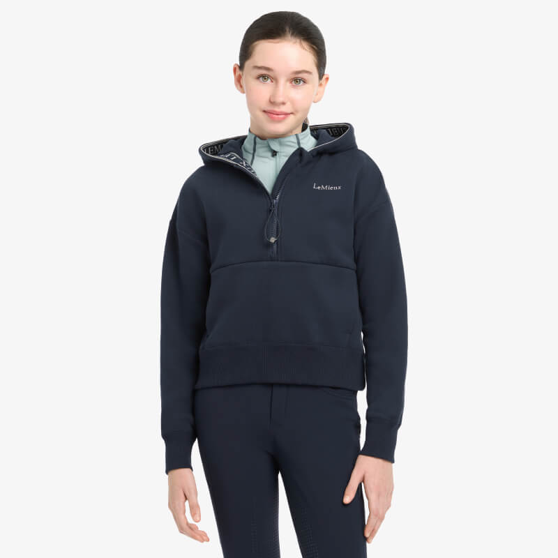 Young rider in LeMieux Hettie Half Zip Hoodie Navy stands outside, background blurred.