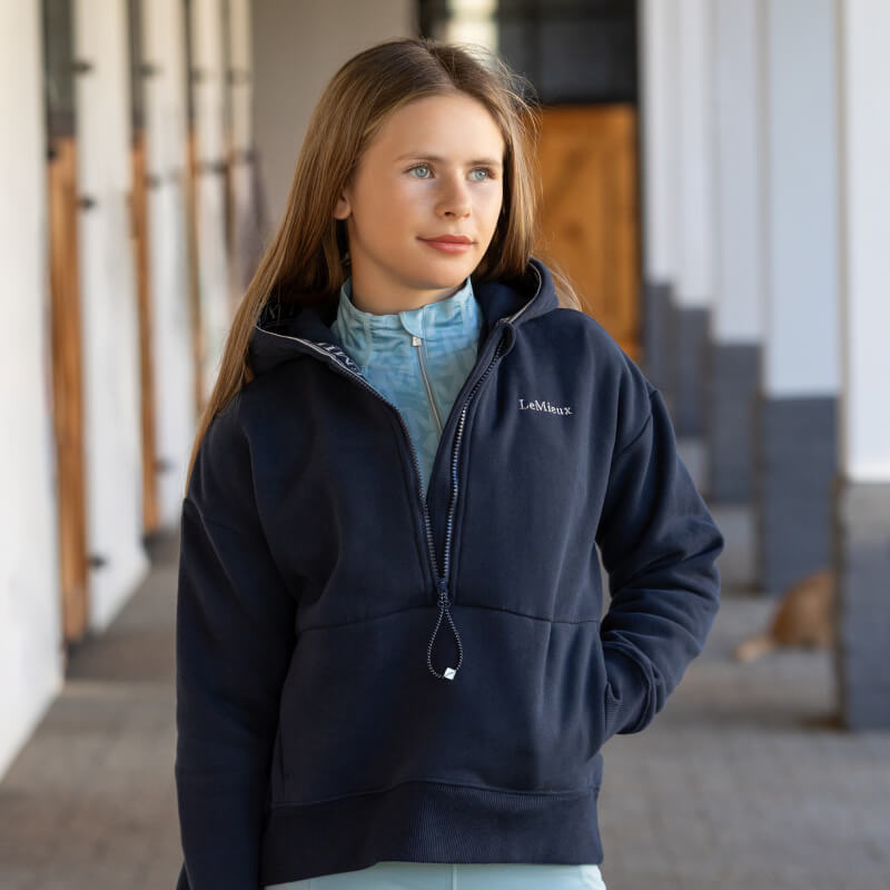 Young woman in a LeMieux Young Rider Hettie Half Zip Hoodie Navy stands outside, looking aside.