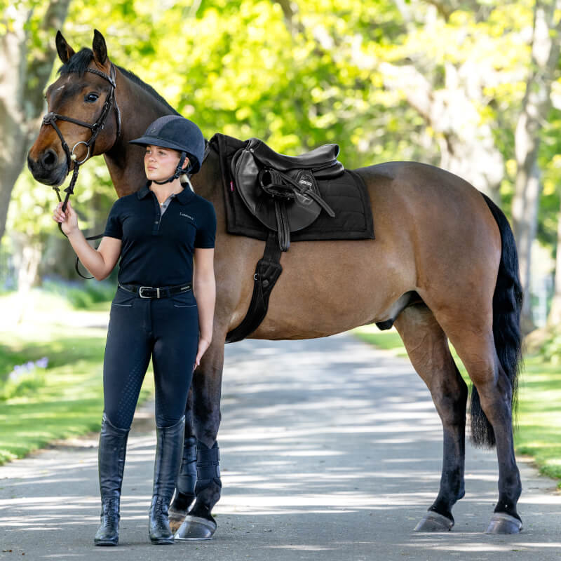 Woman in riding gear stands by a brown horse with a LeMieux Pillow Pad Black saddle pad.