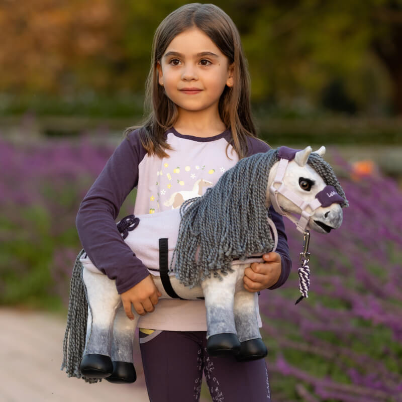 A young girl holds the LeMieux Toy Pony Blanca outside with purple flowers behind her.