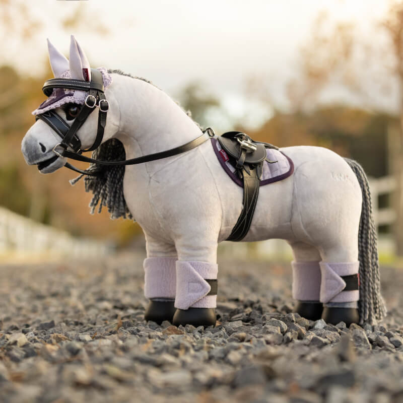 LeMieux Toy Pony Blanca, with bridle, saddle, and leg wraps, stands on a gravel path.