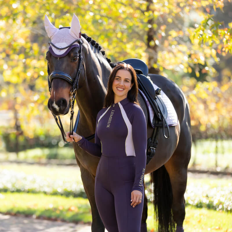 Smiling woman in a LeMieux Phillipa Mesh Baselayer Juniper stands beside a horse outdoors.