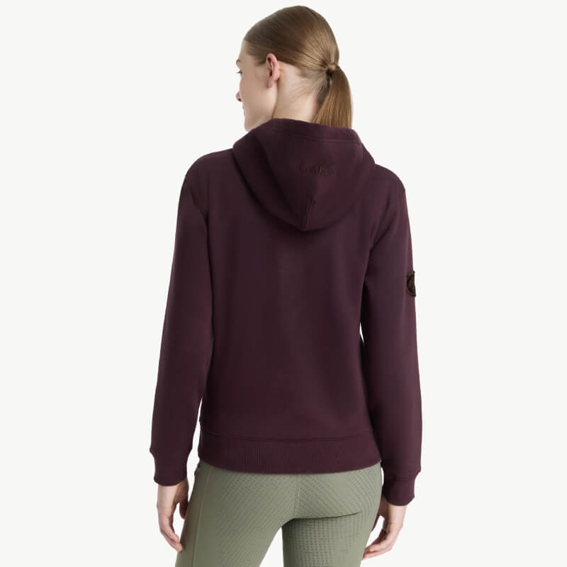 Woman in a LeMieux Elspeth Hoodie Damson and green pants stands facing away from camera.