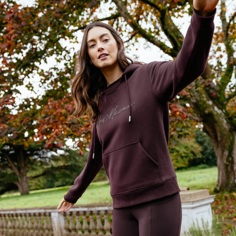 Woman in a LeMieux Elspeth Hoodie Damson enjoys autumn trees outdoors.