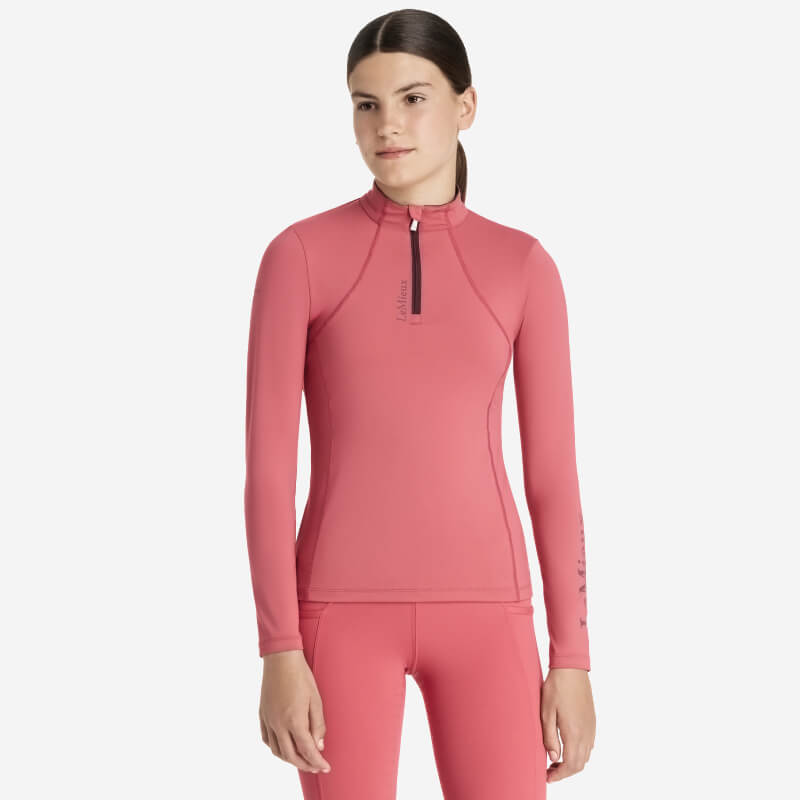 Young rider in LeMieux Young Rider Classique Baselayer Cranberry holding a matching pink helmet.