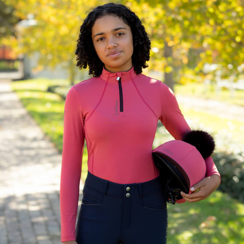 Young rider in LeMieux Young Rider Classique Baselayer Cranberry holding a matching pink helmet.