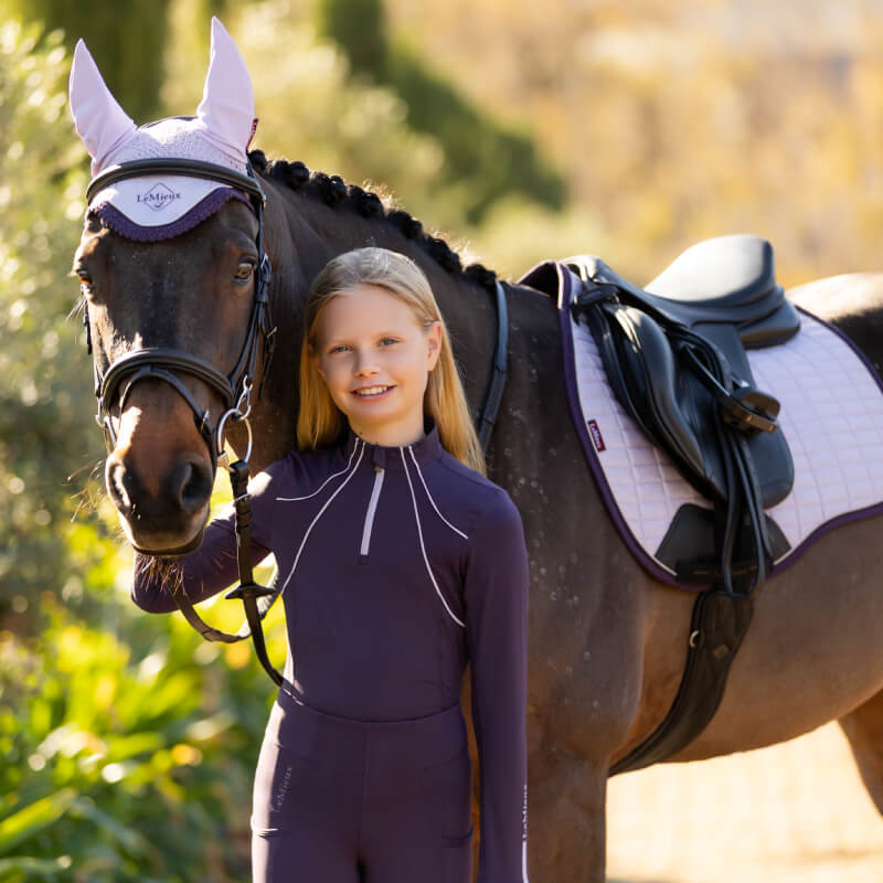 Smiling girl in LeMieux Young Rider Mia Mesh Baselayer Juniper stands by a saddled horse.