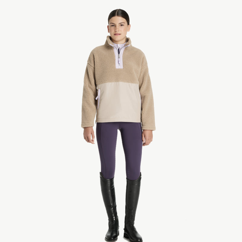 Woman in LeMieux Young Rider Tammy Teddy Fleece Almond, purple leggings, black knee-high boots.