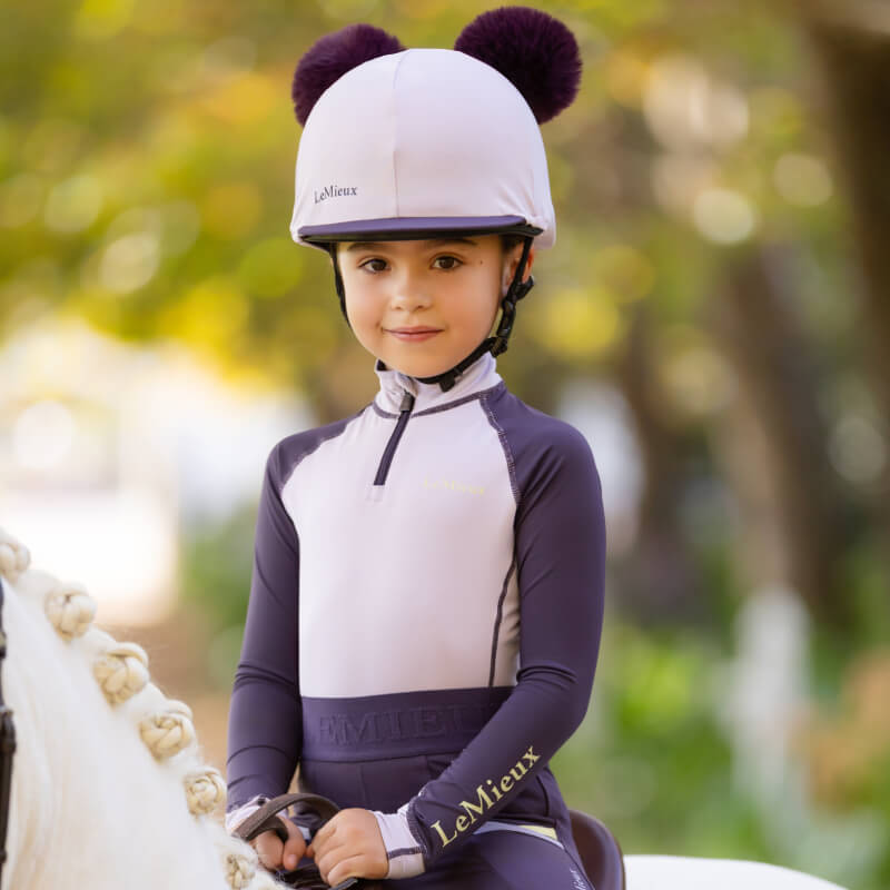 Young girl in LeMieux Mini Baselayer Lilac and helmet sits on a white horse outdoors.