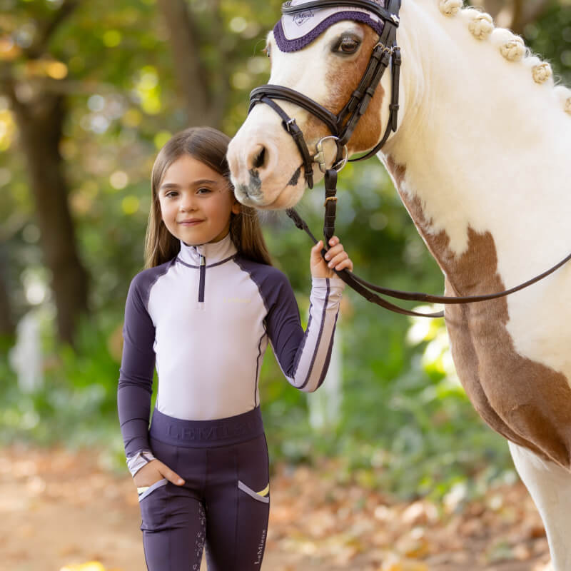 Young girl in a LeMieux Mini Baselayer Lilac stands beside a white and brown horse outdoors.