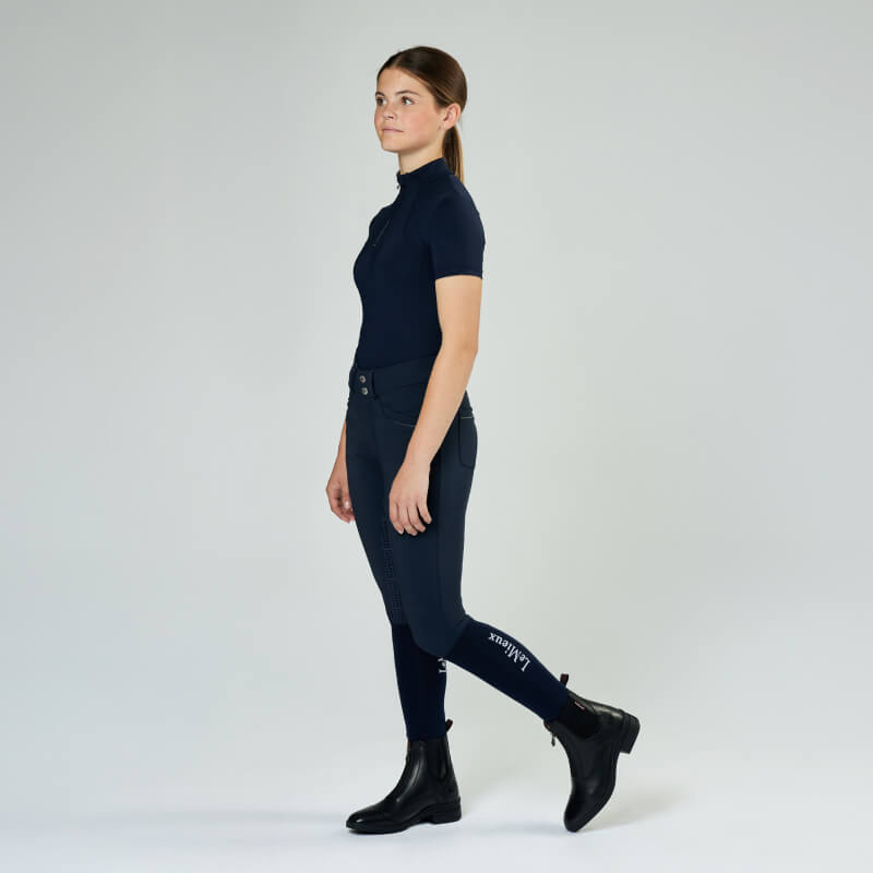 Girl wears LeMieux Junior Zip Paddock Boot Black, standing sideways on a plain light background.