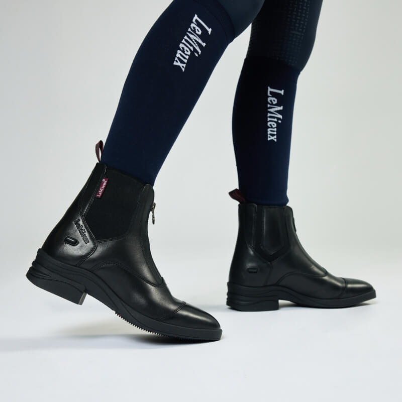 Wearing LeMieux Junior Zip Paddock Boot Black with navy LeMieux riding tights, side view.