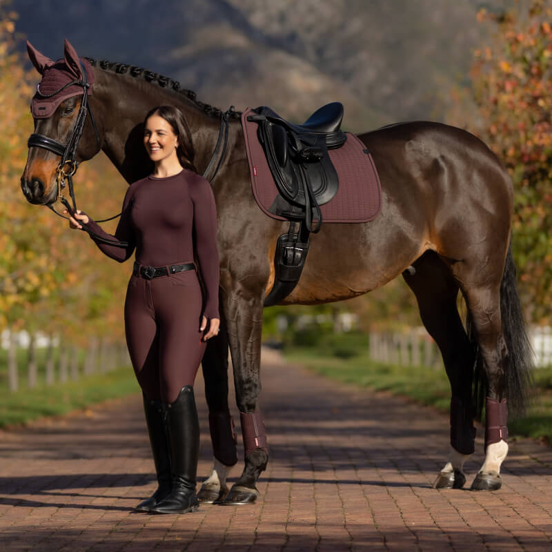 Woman in maroon stands by horse with LeMieux Essence Dressage Square Damson saddle pad.