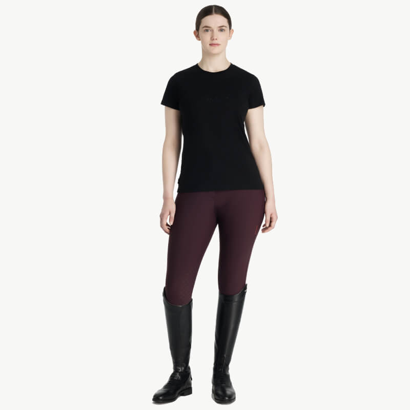 Woman in a LeMieux Classique Sparkle T-Shirt Black, burgundy pants, and tall black boots.