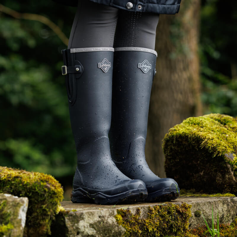 Person wearing LeMieux Ultra Stride Wellington Boots Navy stands on mossy rocks outdoors.