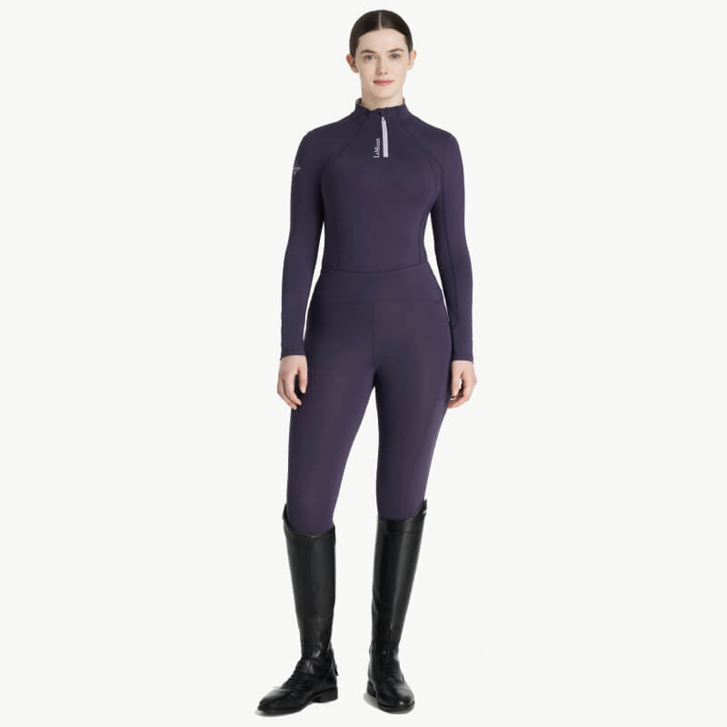 Woman in a LeMieux Classique Baselayer Juniper and black riding boots, facing forward.