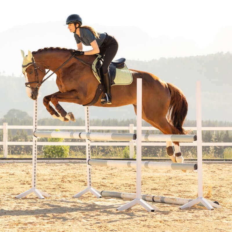 A rider on a LeMieux Suede Close Contact Square Macaron clears an outdoor jump.