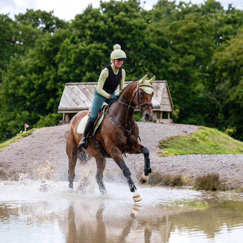 Rider on horseback crosses shallow water, horse in LeMieux Suede Close Contact Square Macaron.