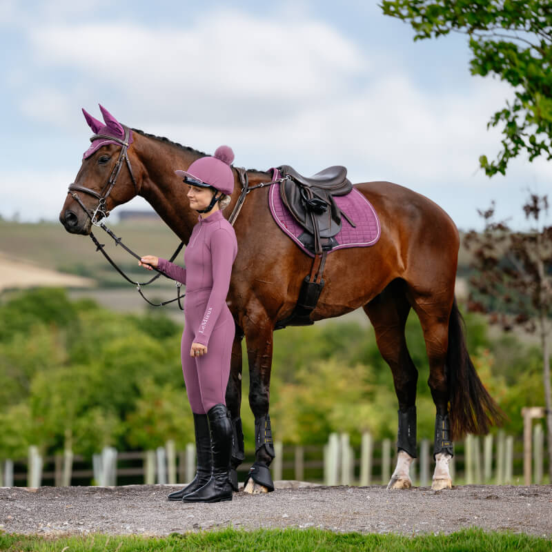 Rider in mauve stands by brown horse with a LeMieux Suede Close Contact Square in Mallow.