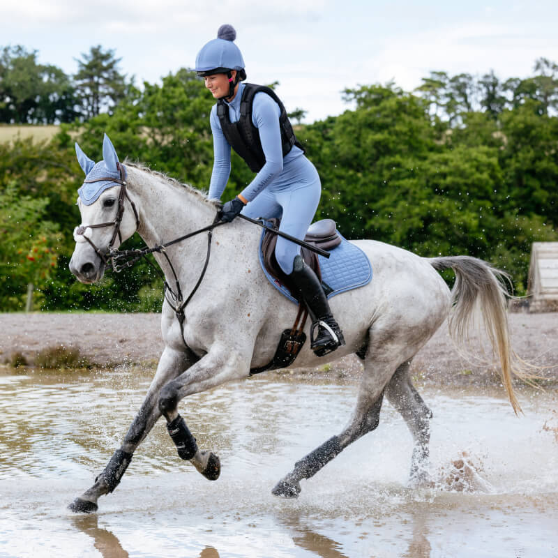 A rider on a gray horse crosses water with the LeMieux Suede Close Contact Square in Powder Blue.