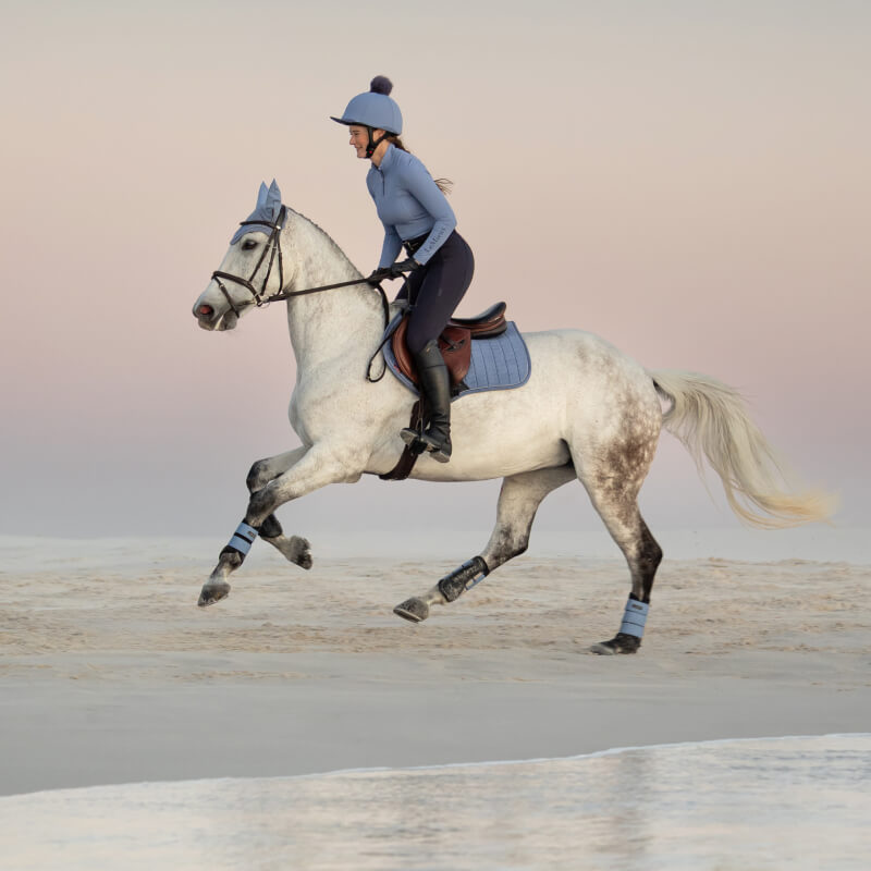 Rider in blue with LeMieux Suede Close Contact Square Powder Blue on gray horse at sunset beach.