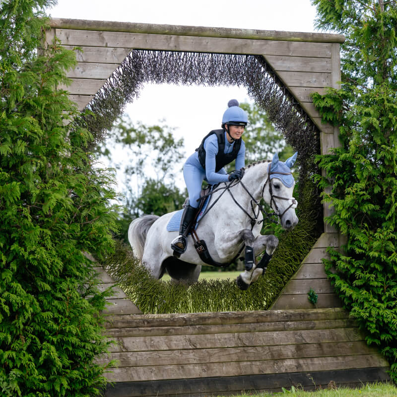 A rider in blue jumps a gray horse with a LeMieux Suede Close Contact Square Powder Blue.