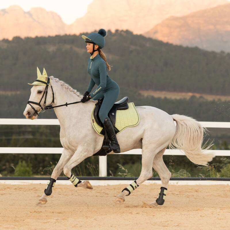 Woman in green rides a white horse with a LeMieux Suede General Purpose Square Macaron pad.