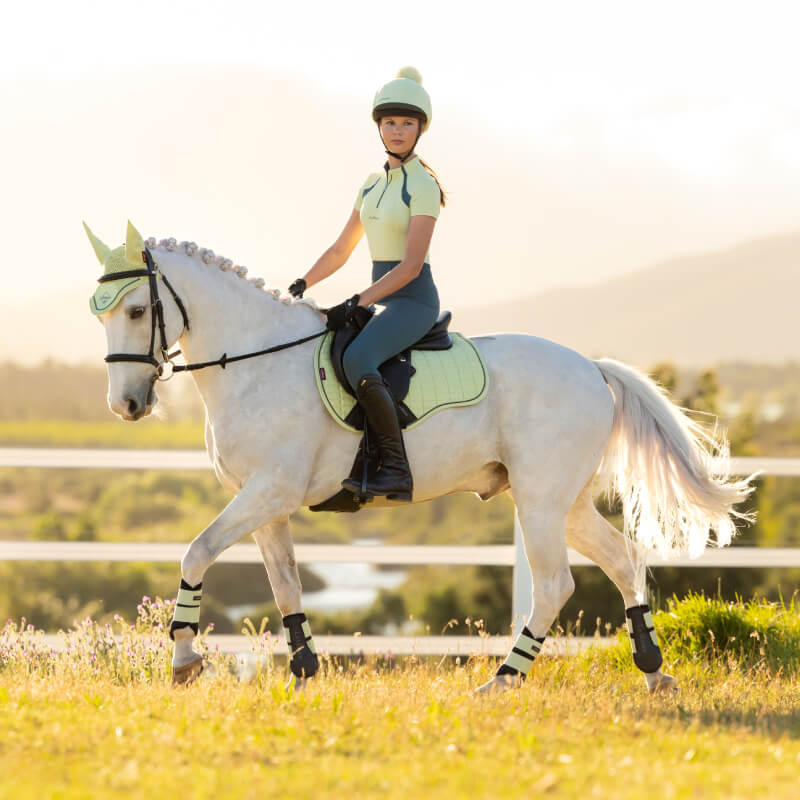 Woman in yellow rides a white horse with a LeMieux Suede GP Square Macaron pad in a sunny field.