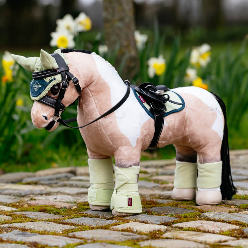 A plush toy horse with a LeMieux Toy Pony Pad Macaron stands on cobblestones, flowers behind.