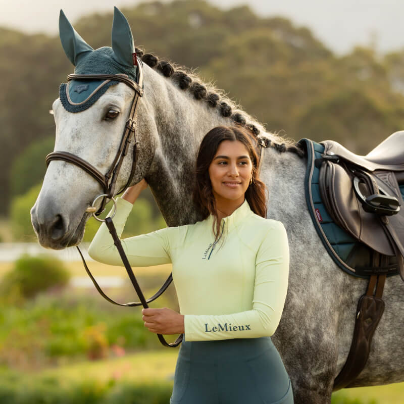 Woman in LeMieux Classique Baselayer Macaron stands by a gray horse with braided mane outdoors.