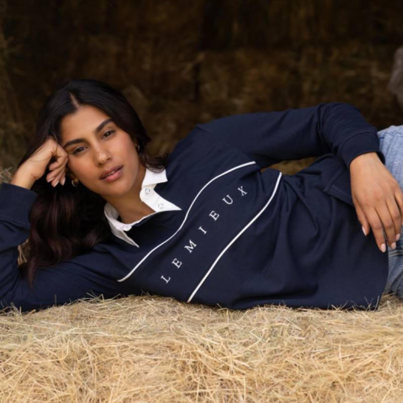 Woman in LeMieux Rochelle Rugby Shirt Navy lies on hay, facing camera.