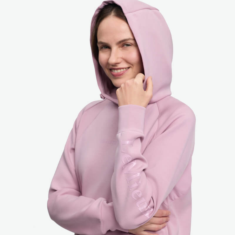 Smiling woman in a LeMieux Nadine Airflow Hoodie Fondant, hand near chin, plain background.