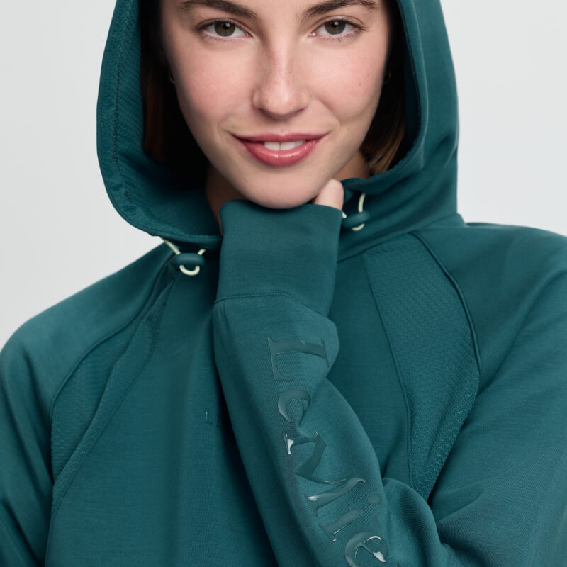 Smiling woman in LeMieux Nadine Airflow Hoodie Jungle, hand near chin, white background.