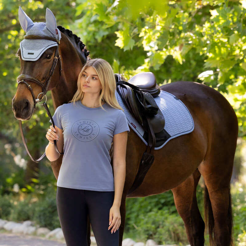 Woman in LeMieux Classique T-Shirt Powder Blue stands by a saddled horse beneath leafy trees.