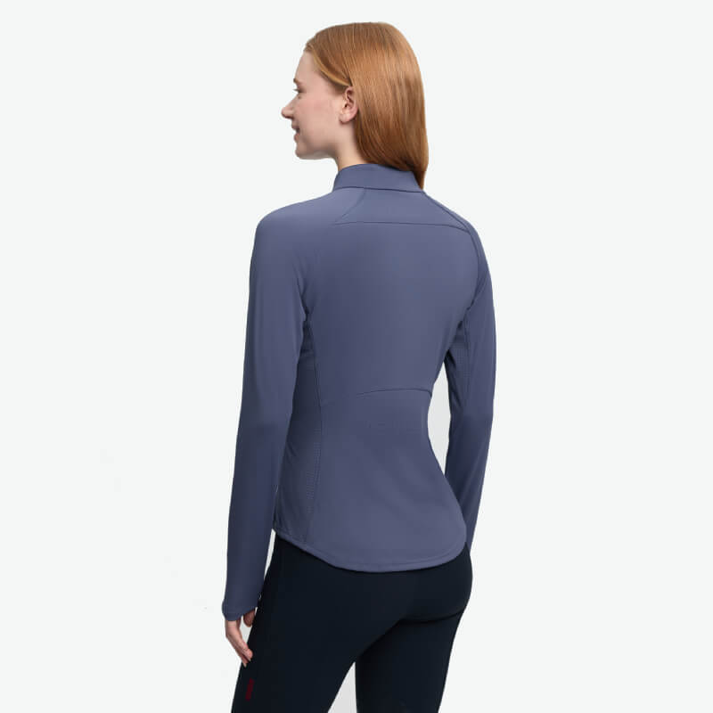 Woman with long hair wears a blue LeMieux Airflow Long Sleeve Baselayer Shadow, back view.