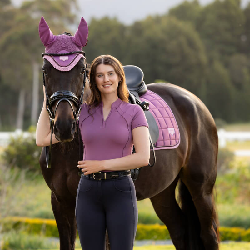 Woman wears LeMieux Lisa Mesh Baselayer Mallow, standing by horse with matching purple tack outside.