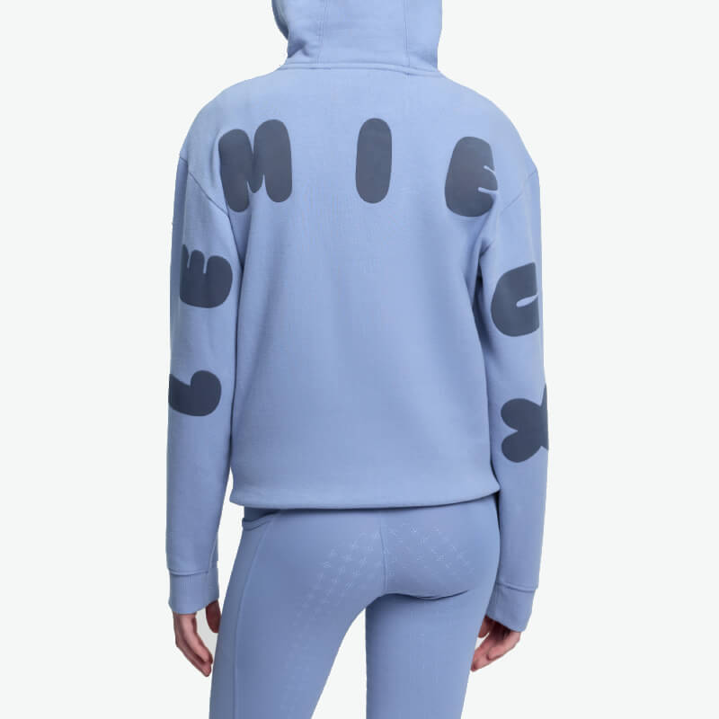 Smiling woman in LeMieux Young Rider Sia Hoodie Powder Blue, white background.