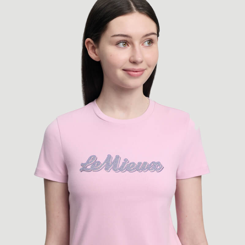 Woman wears LeMieux Young Rider Arianna T-Shirt Fondant and blue leggings against plain background.