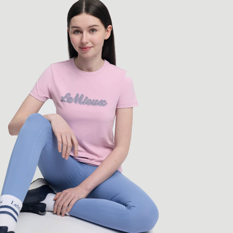 Smiling woman in a pink LeMieux Young Rider Arianna T-Shirt Fondant and blue leggings sits.