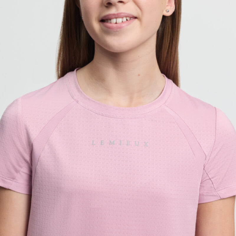 Teen girl in LeMieux Young Rider Sports T-Shirt Fondant and black pants, smiling by a white wall.