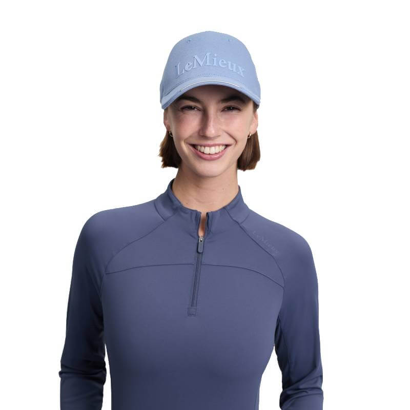 Smiling person in blue long-sleeve shirt and LeMieux Lara Cap Powder Blue One Size on white.