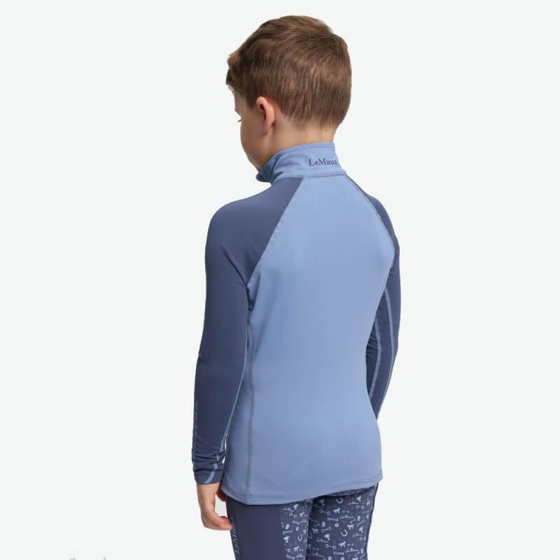 Young boy in LeMieux Mini Baselayer Powder Blue, standing and facing away in blue long sleeves.