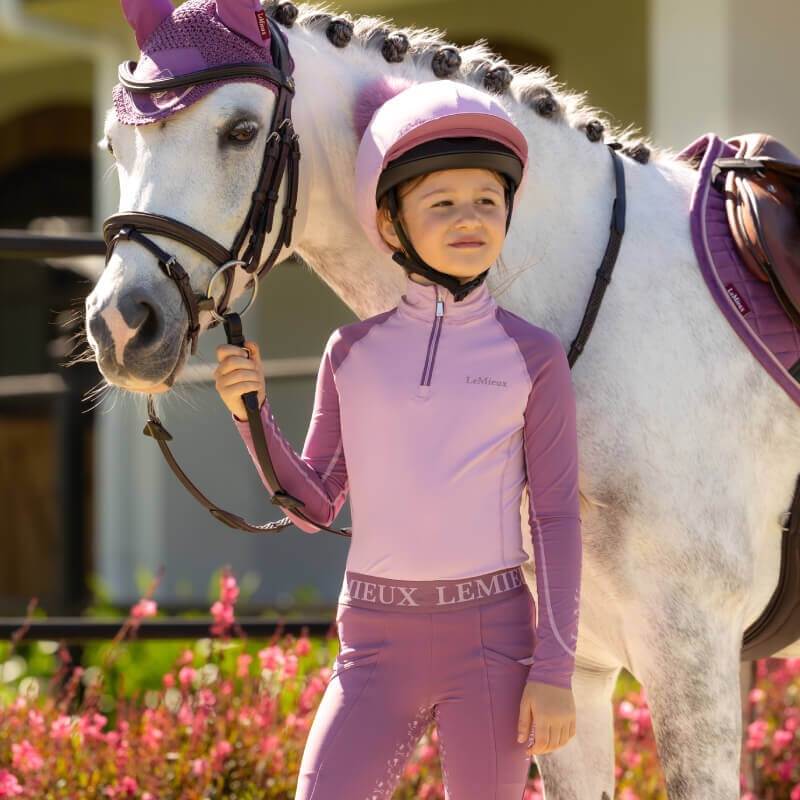 Child in LeMieux Mini Baselayer Fondant and purple gear stands by white horse with purple tack.
