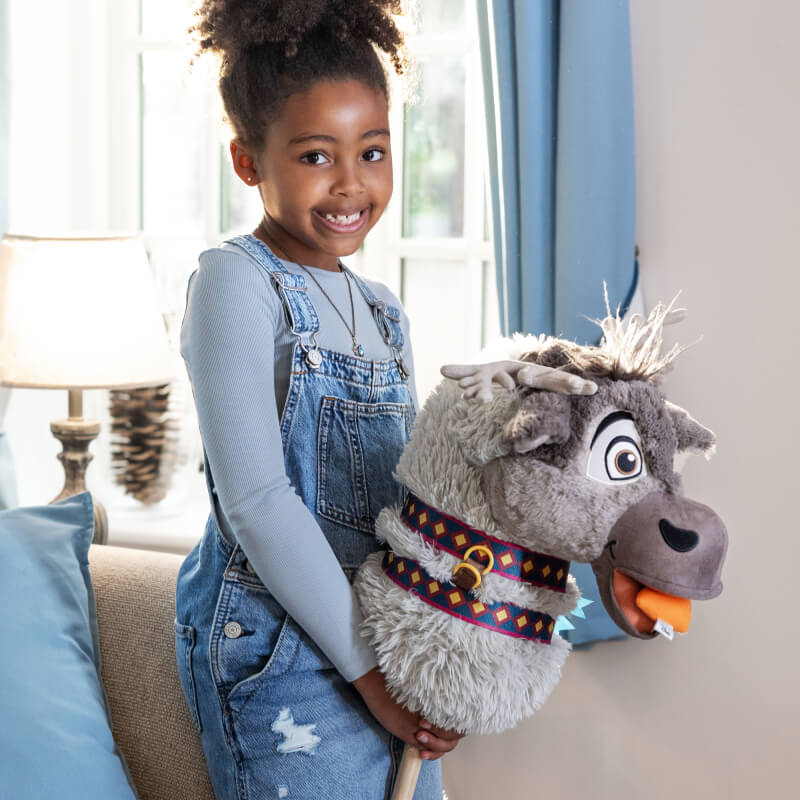 Girl in overalls holds a LeMieux Disney Hobby Horse Sven toy indoors by a window.