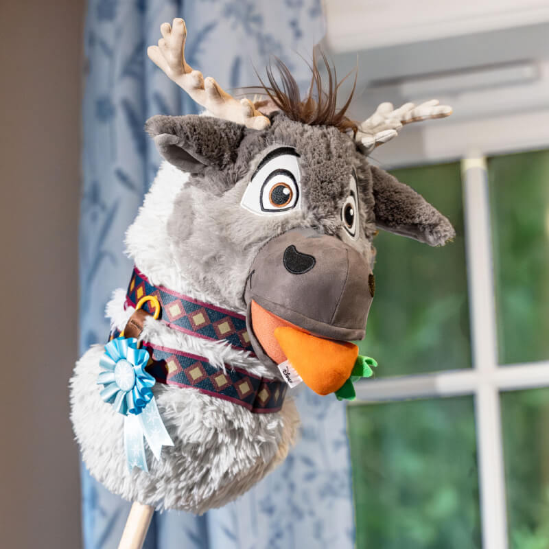 A LeMieux Disney Hobby Horse Sven stands indoors by a window with blue curtains and ribbon.