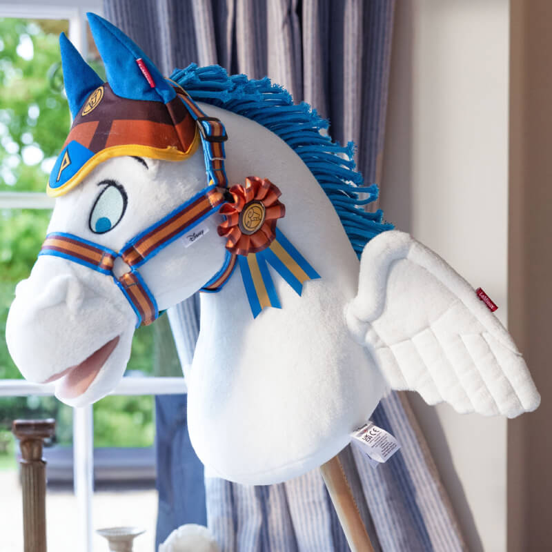 LeMieux Disney Hobby Horse with Pegasus accessories, blue wings, and ribbon in a sunlit room.