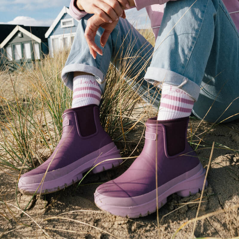 Person in LeMieux Bramshaw Chelsea Welly Mallow and striped socks sits on sand near houses.